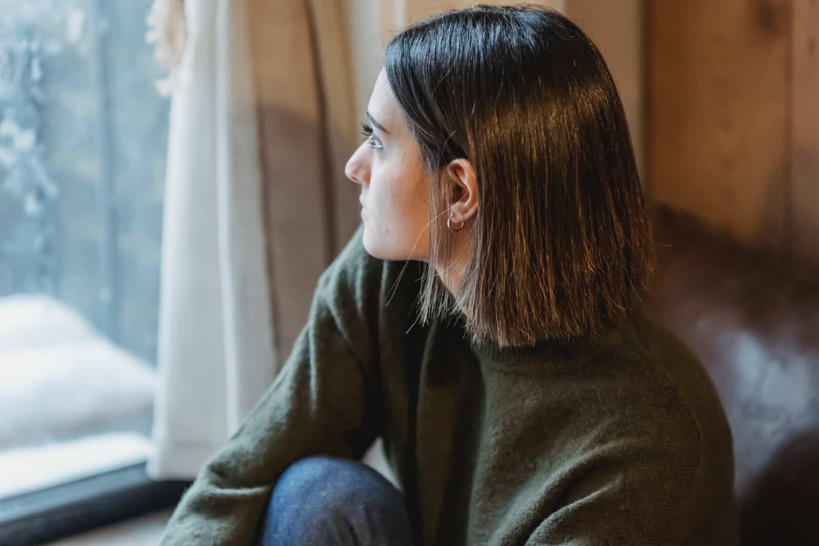 mujer pensativa en suéter marrón mirando por la ventana
